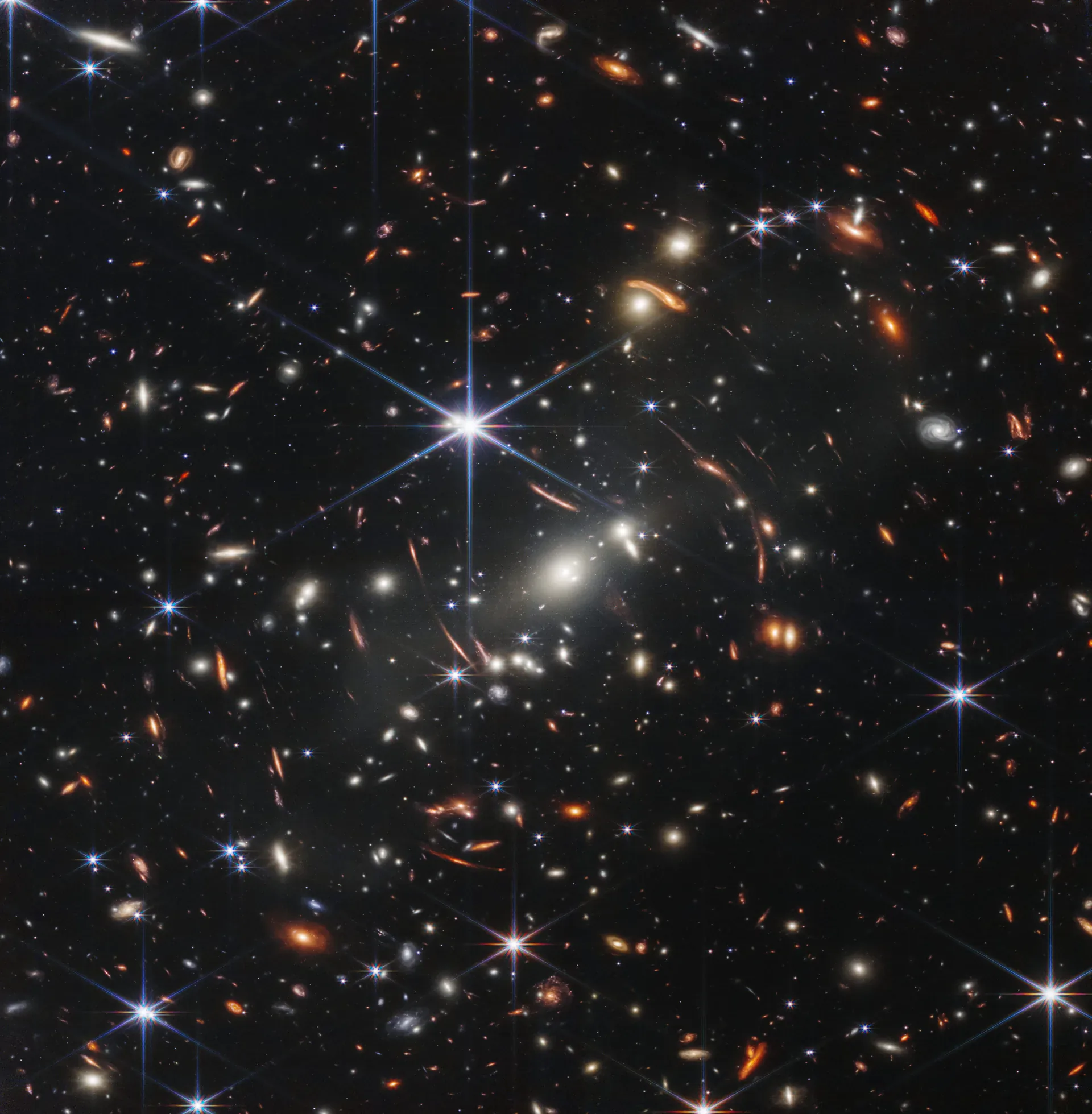 Primer campo profundo de Webb, con numerosas galaxias y estrellas sobre el espacio negro, picos de difracción alrededor de estrellas brillantes y galaxias rojas deformadas por lentes gravitatorias